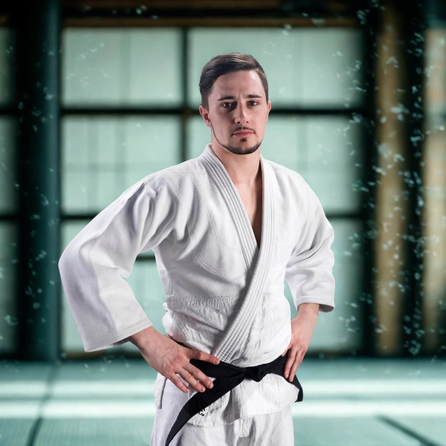 Man in a white martial arts KROLIN PureGi Judo Uniform – 450gsm cotton jacket and poly-cotton pants a black belt standing in a dojo.