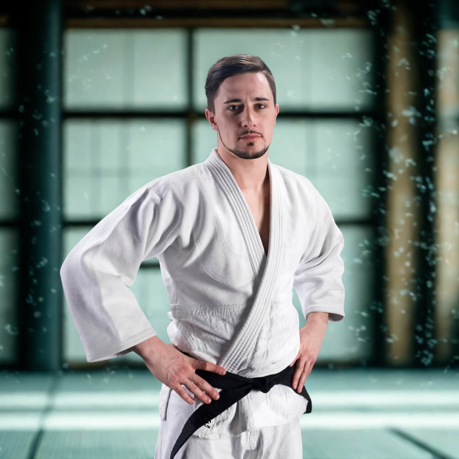 Man in a white martial arts KROLIN PureGi Judo Uniform – 450gsm cotton jacket and poly-cotton pants  a black belt standing in a dojo.